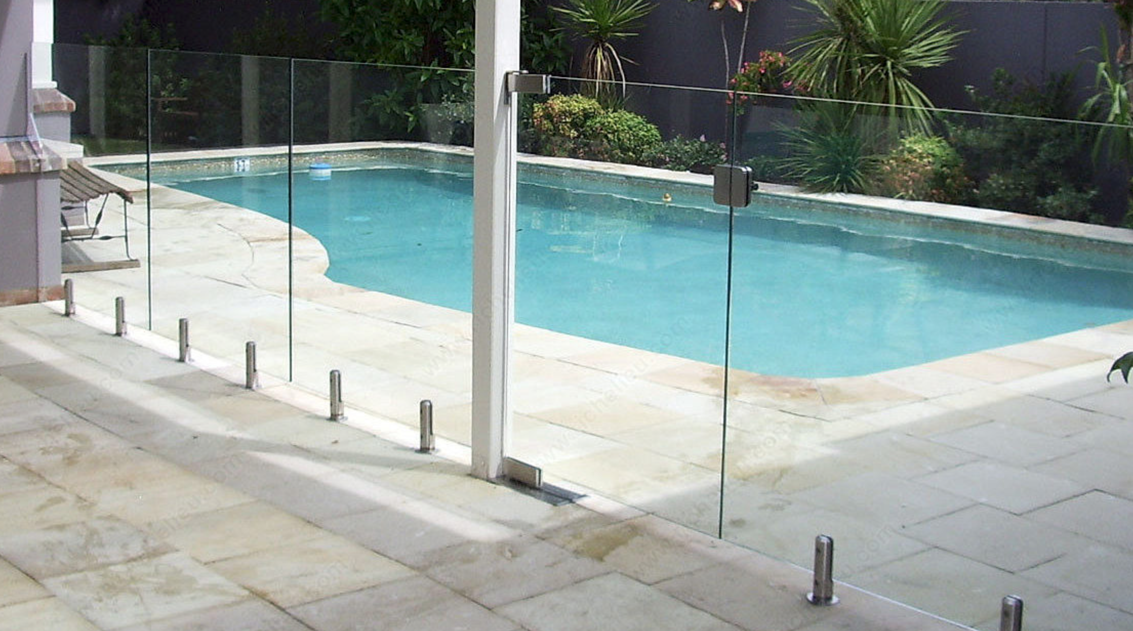 Garde-corps en verre transparent autour d’une piscine extérieure, fixations métalliques et rails de sécurité, jardin et terrasse pavée en arrière-plan.